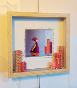 tableau vitrine décorative à partir d’une reproduction de son petit personnage appelé Poupir et décoré d’éléments de customisation composé de rubans perles origami